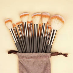 A set of makeup brushes with black and silver striped handles and cream bristles with orange tips, arranged in a brown drawstring pouch against a warm beige background.
