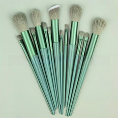 A set of makeup brushes with metallic sage green handles and light beige bristles, arranged in a fan shape against a solid pale green background.