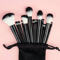 A 10-piece makeup brush set with black handles and black-and-white bristles, arranged in a fan shape above a black drawstring pouch on a solid pink background.