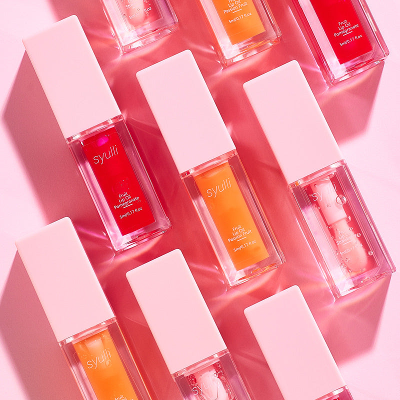 Seven Syulli lip gloss bottles arranged diagonally on a pink background. The clear rectangular bottles show shades of red, orange, and pink gloss inside, with white square caps.