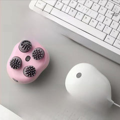 A pink handheld facial massage device with four black textured massage heads, shown on a light gray desk next to a white computer mouse and keyboard.