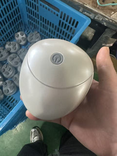A person's hand holding a small, rounded, egg-shaped white electronic device with a power button symbol on top, set against a background of a blue plastic crate and green floor.